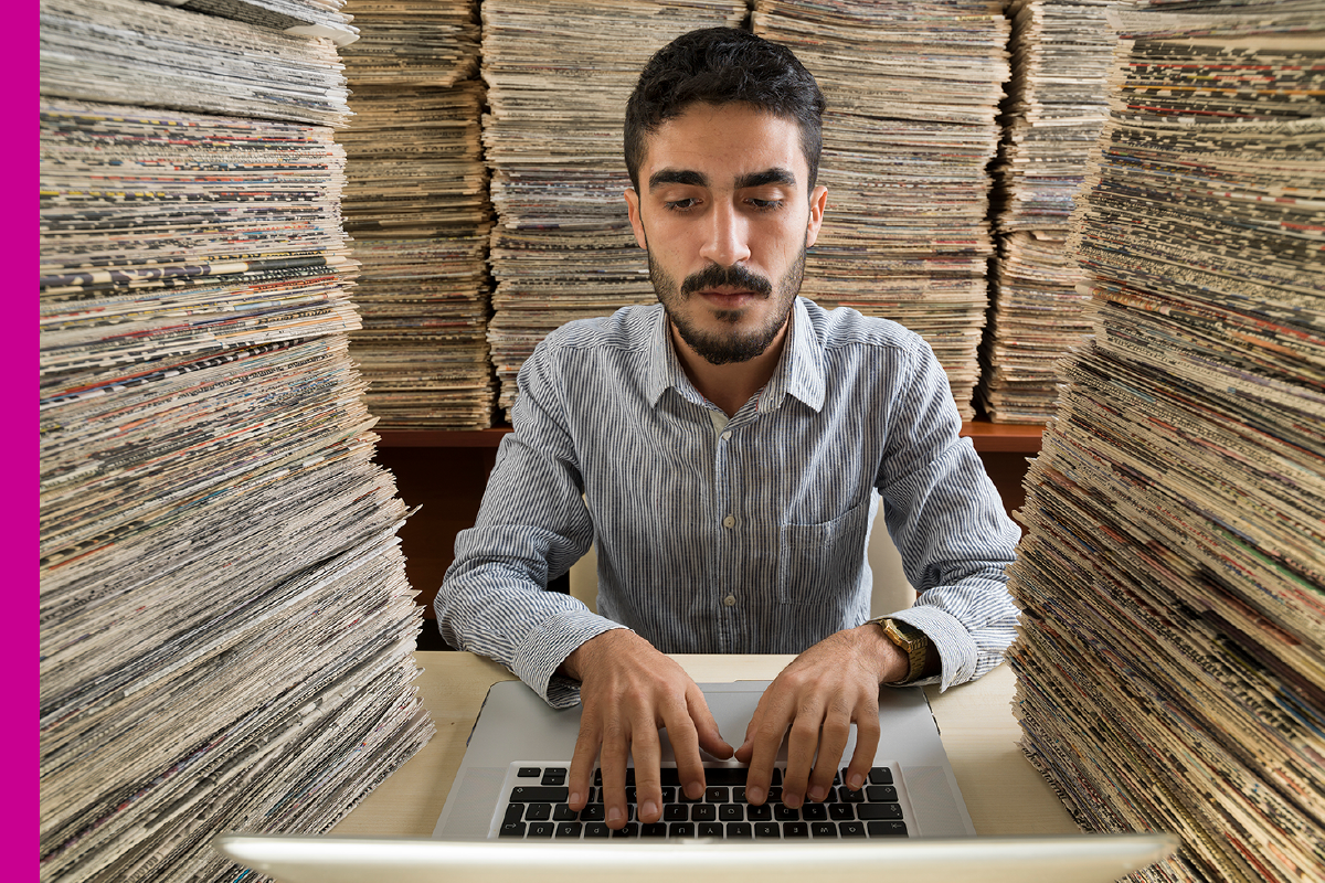 A man typing on a laptop, surrounded by a stack of files.