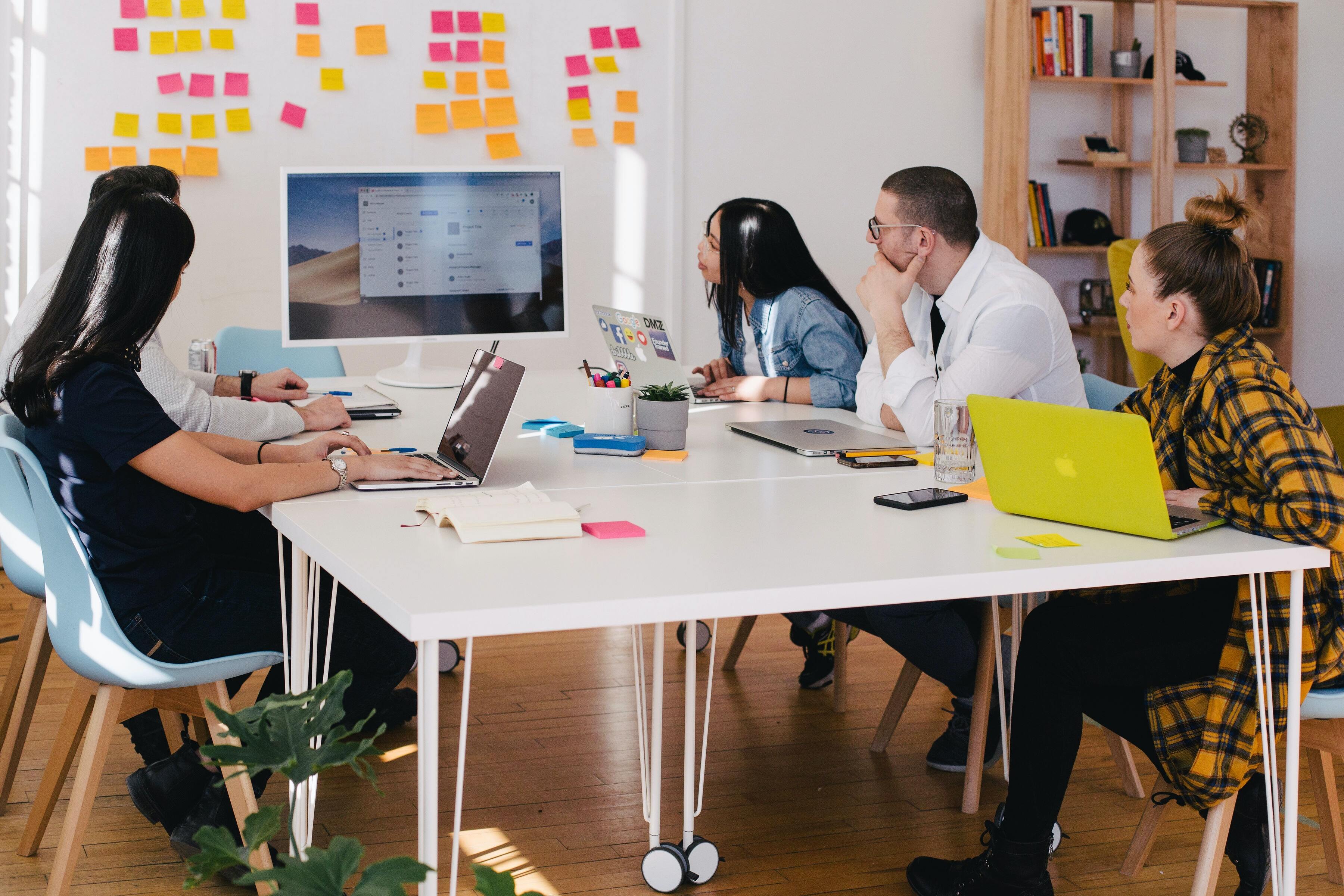 A group of five people sitting around a desk with their laptops looking at a board and planning.