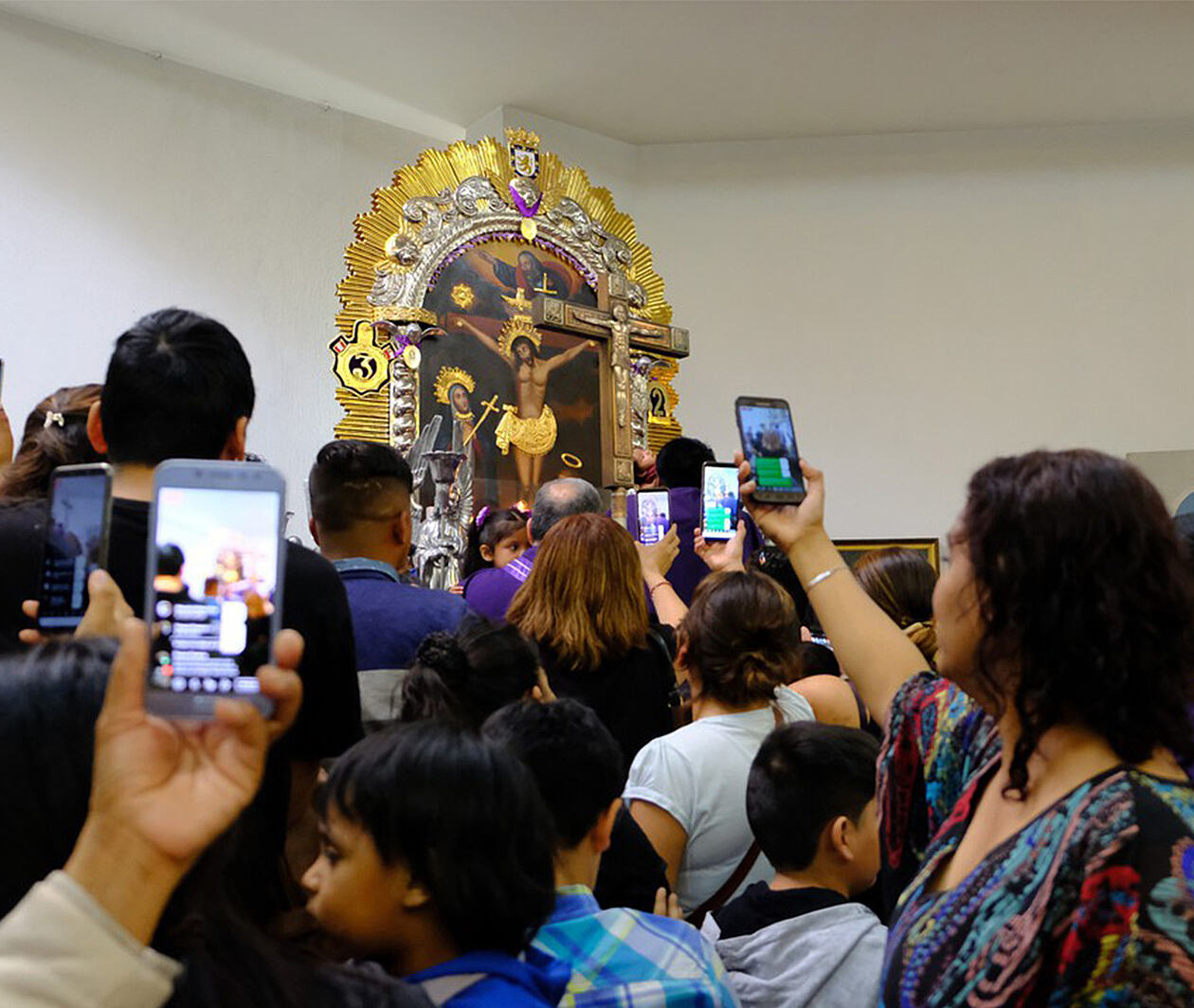 Interior of a church with a painting of the Christ, surrounded by people aiming their smartphones at the image. These are Peruvian migrants honoring in Chile the Lord of Miracles, the most revered Peruvian religious icon. 