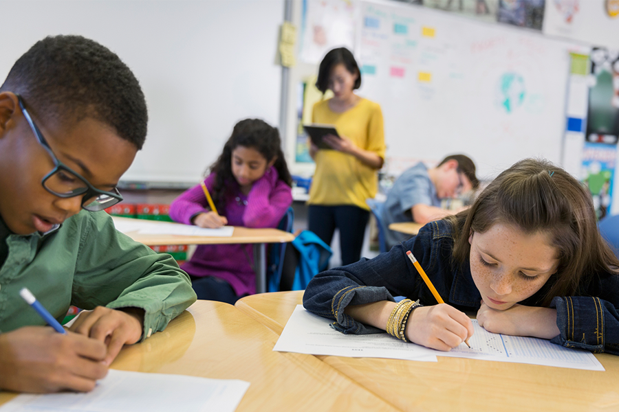 Students seated at desks in a classroom, concentrate on completing their exam papers while a teacher stands nearby.