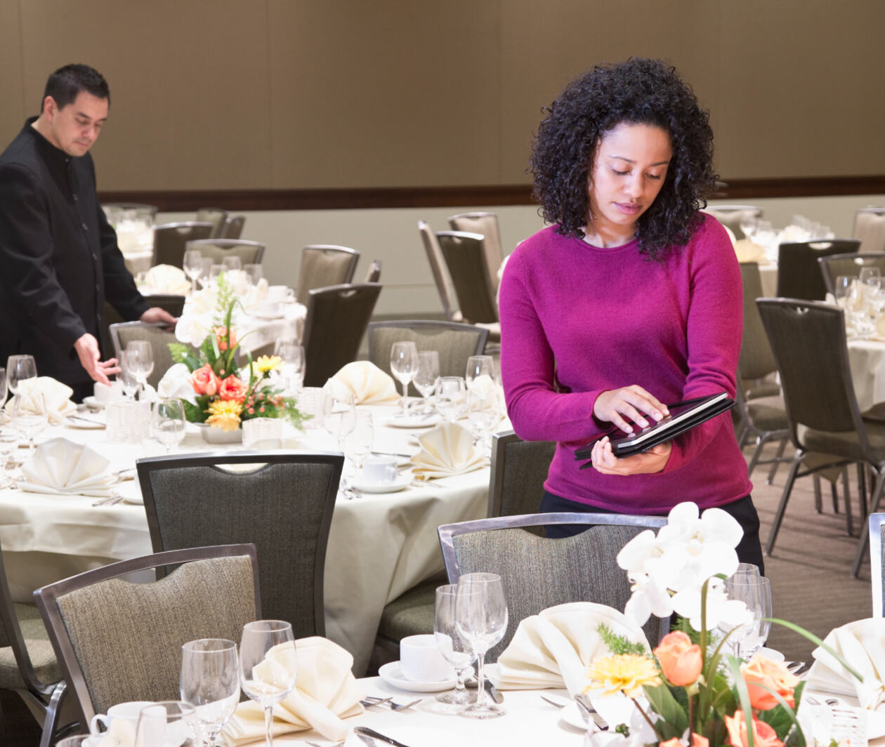 Woman in a function room, surrounded by round tables decorated for a banquet or event, holding an ipad. 