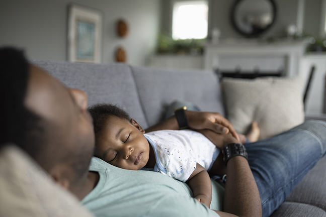 Tired baby son sleeping on father on living room sofa